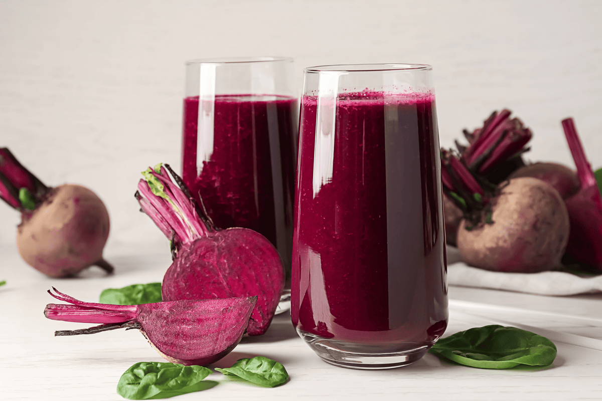 A glass of beet juice surrounded by fresh beetroot with few green leaves on a white table with benefits