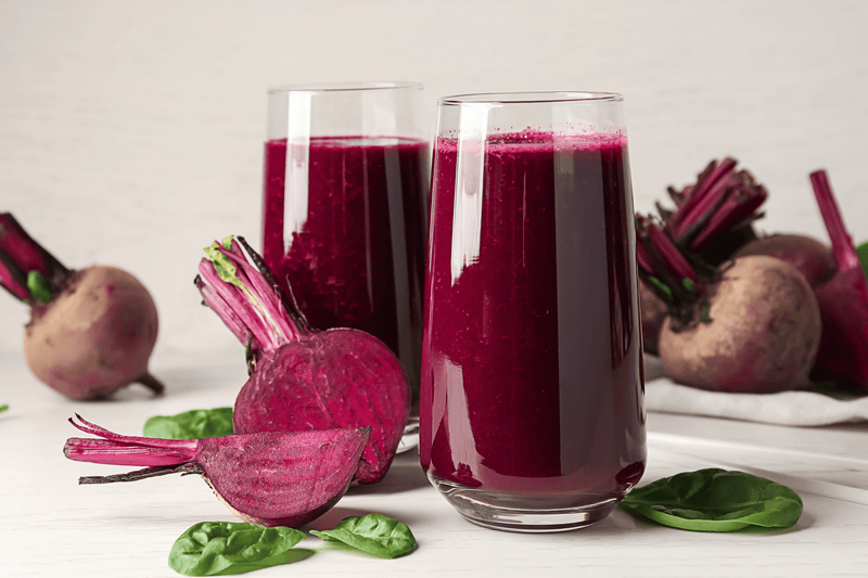 A glass of beet juice surrounded by fresh beetroot and spinach leaves on a wooden table.