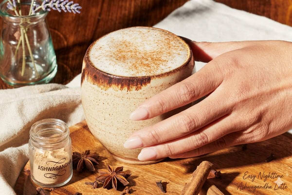 A warm ashwagandha latte in a ceramic mug surrounded by cinnamon sticks, cardamom, and turmeric powder on a wooden surface