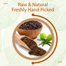 Raw, freshly hand-picked black pepper powder spilling from a wooden scoop beside a bowl of whole black peppercorns, fresh pepper vine sprigs, and bright green leaves on a white background.