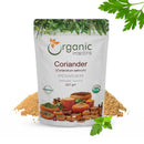 Organic Mantra Coriander Powder 227g standup pouch (Coriandrum sativum) with USDA Organic and OneCert certifications, origin India, flanked by fresh coriander herb sprigs and a mound of ground powder.