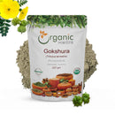 Organic Mantra Gokshura Tribulus terrestris Powder, 227g white resealable standing pouch with USDA Organic and OneCert Organic certifications and India origin label, centered on a white background with a grey-green powder splash behind it, bright yellow. Gokshura flowers and spiky raw fruit cluster at the top left and a single spiked Tribulus terrestris fruit at the bottom right.